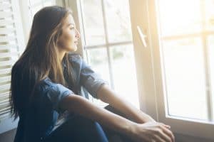 Young woman sitting and looking through window Smiling young woman sitting and looking through the window. With long hair, wears denim shirt. Hands on legs.